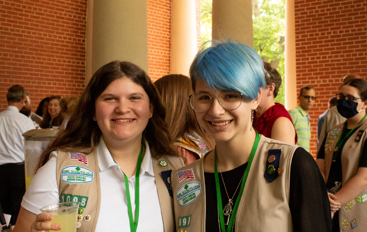 two older girl scouts, one with long brown hair and one with short blue hair, pose together outside the venue at the Highest Awards ceremony.