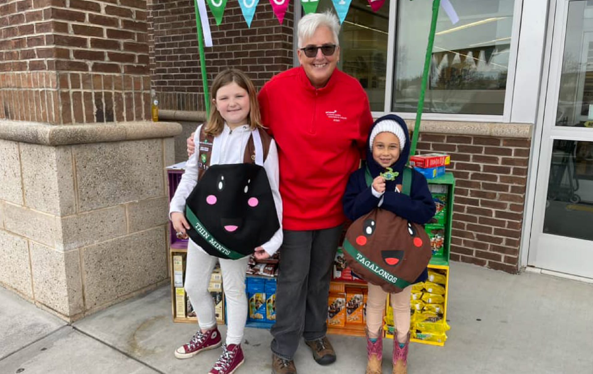 CEO Lora poses with Girl Scouts dressed as a Thin Mint and a Tagalong at a booth.