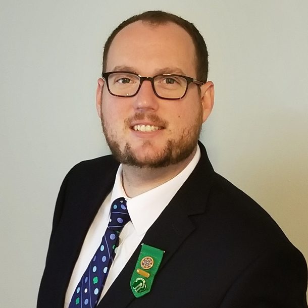 Chris bullard is a person with very short brown hair and is wearing glasses, a navy jacket, and a girl scout tie