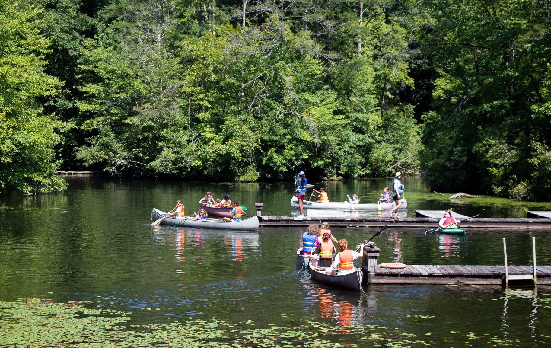  several Girl Scout campers row gray canoes around the Camp WaBak lake 