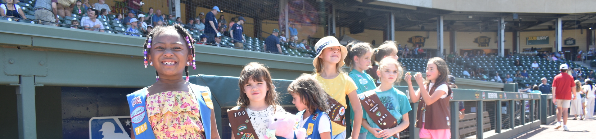  A group of Girl Scouts in uniform smile at Greenville Drive mascot doing a split on the field during Girl Scout Day at the Greenville Drive. 