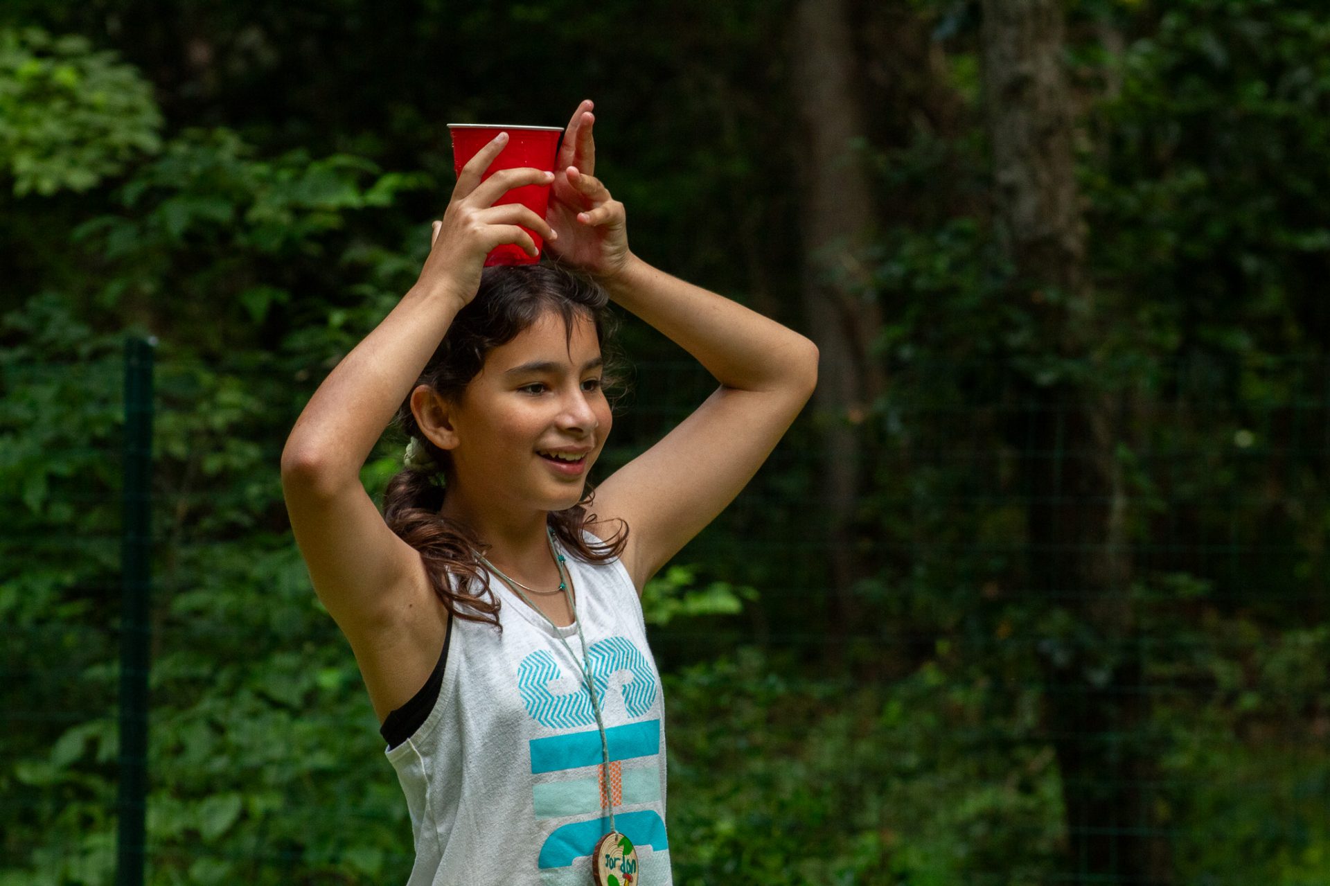 camper in a white tank top with blue words carries a red solo cup on top of her head near trees