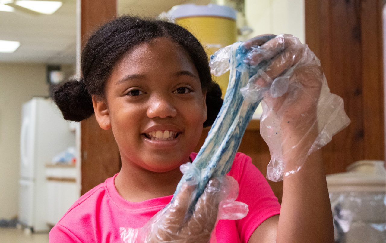 a camper in a pink shirt holds up blue slime