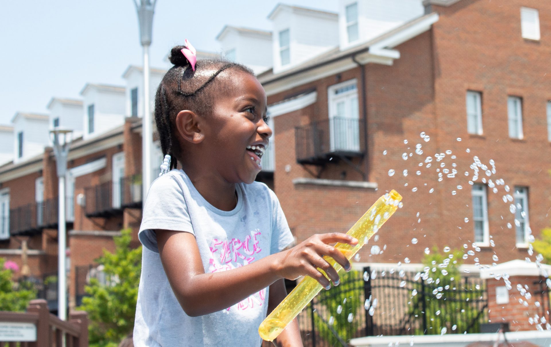 Girl in gray shirt laughs while playing with yellow tube at water fountain