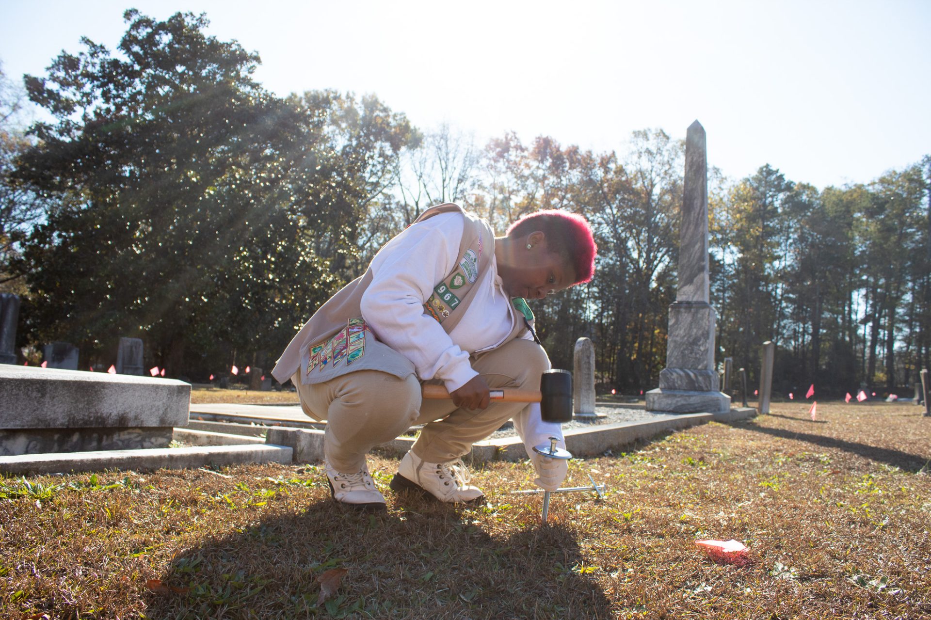  girl scout cadette with short red and black hair in her tan vest hammers a stake into the ground at a cemetery as part of her silver award project. 