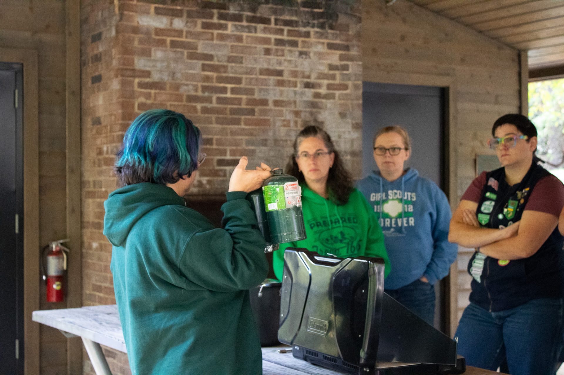  A volunteer with blue hair and a teal hoodie holds a fuel canister in front other volunteers at a outdoor cooking training. 